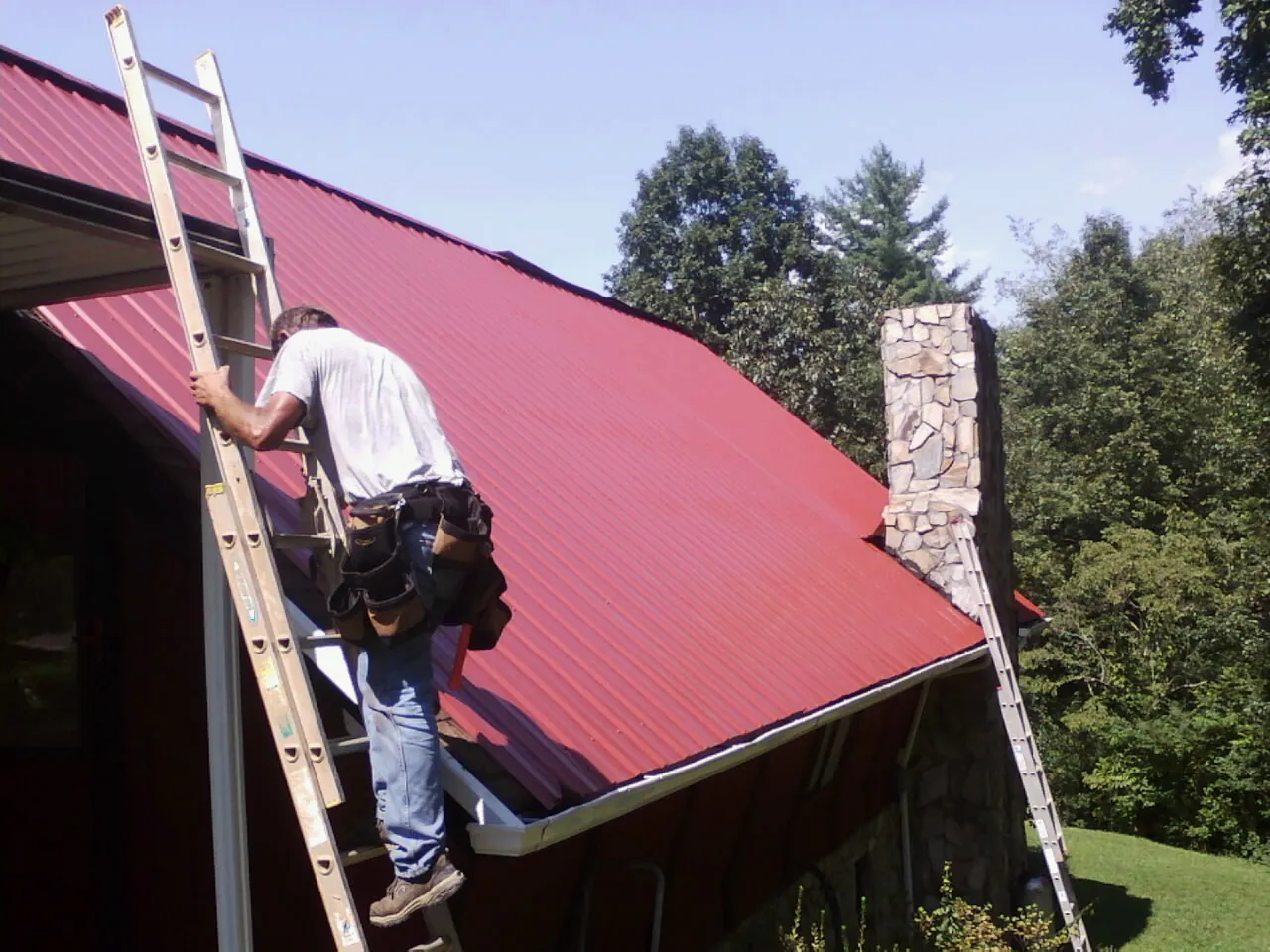 Roofer on a ladder beside a red metal roof and stone chimney — Soffit & Fascia Repair by Roofing Brothers in Fosston